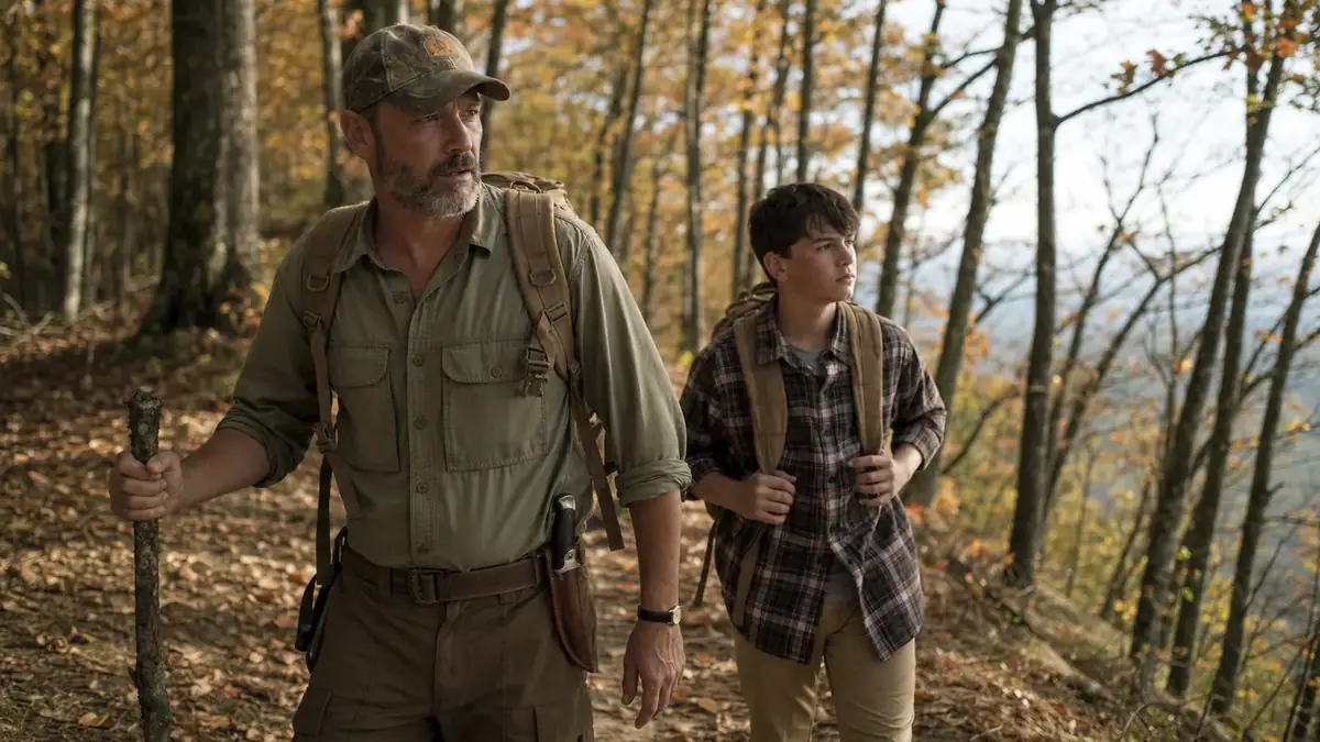 Father and son walking a wooded ridge trail at midday, knife sheathed on the son's belt